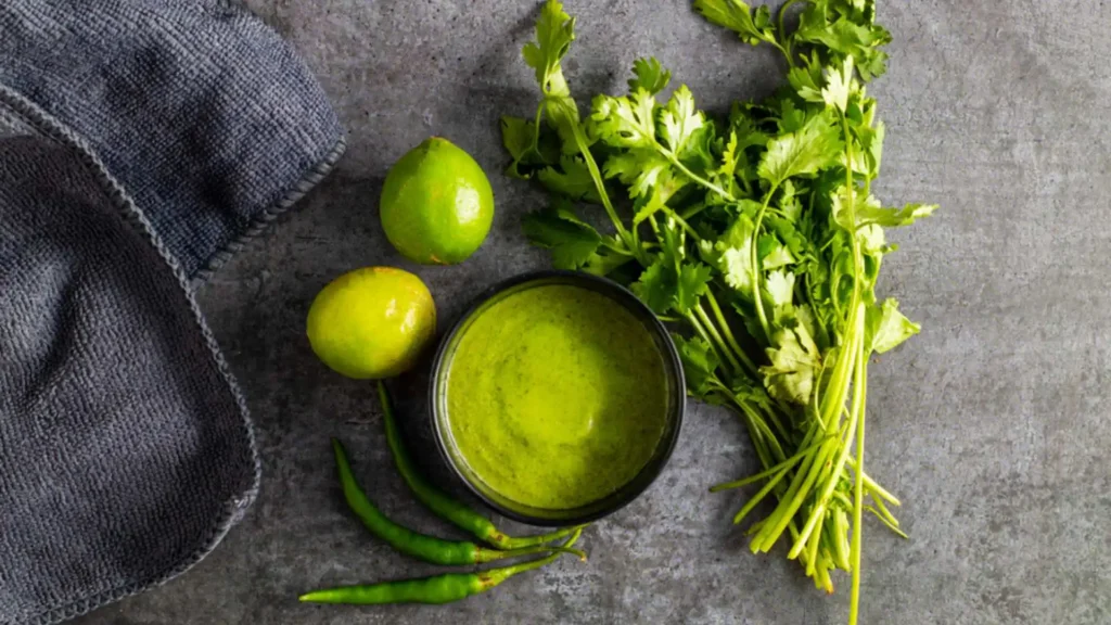 Green mint chutney with limes, cilantro and chili peppers on a dark surface.