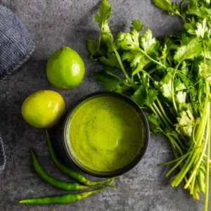 Green mint chutney with limes, cilantro and chili peppers on a dark surface.