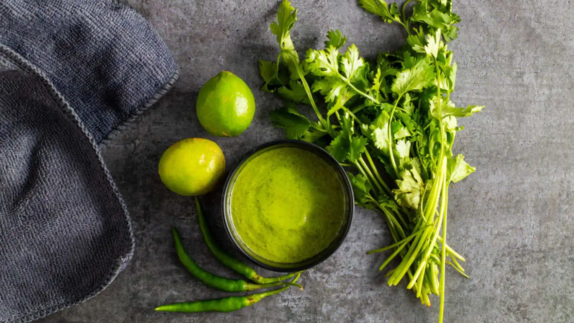 Green mint chutney with limes, cilantro and chili peppers on a dark surface.