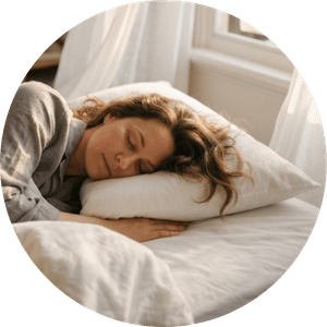 A woman sleeps peacefully in soft morning light with relaxed features and sunlight streaming across the bed, creating a feeling of deep rest, safety, and restoration.
